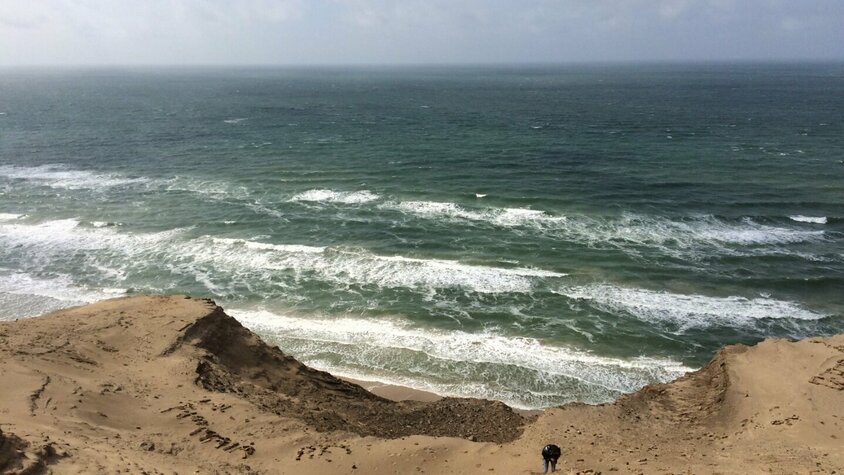 Sanddüne an der dänischen Küste Person erklimmt eine steile Sanddüne an der dänischen Küste mit Blick auf die raue See und brechende Wellen unter bewölktem Himmel.