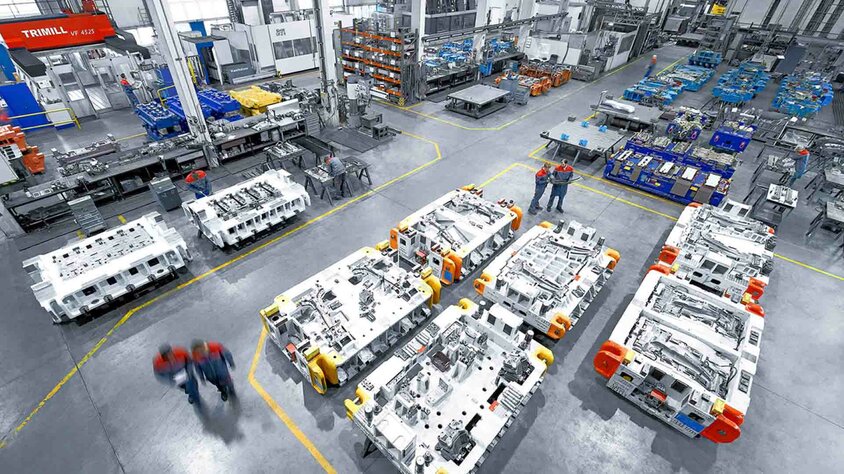 A modern tool manufacturing facility with large stamping and pressing tools. The production hall is clean and well-organized, with several large tools placed on the floor. In the background, various machines and workbenches can be seen, while in the foreground, two employees in work uniforms are engaged in conversation. This image showcases the professional and efficient use of tools and machinery in the toolmaking industry, specifically for the automotive sector.