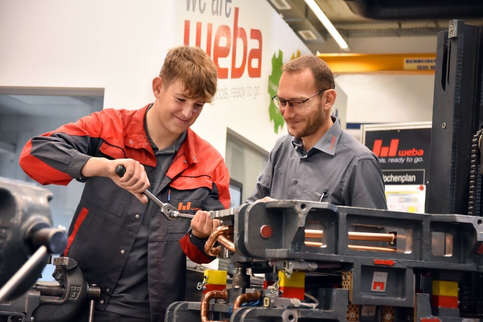 Ein Lehrling in der Werkzeugbautechnik arbeitet an einer Miniatur eines Umformwerkzeugs, während der Ausbilder aufmerksam zuschaut. weba Werkzeugbau bietet Lehre in Steyr in der Metalltechnik an.