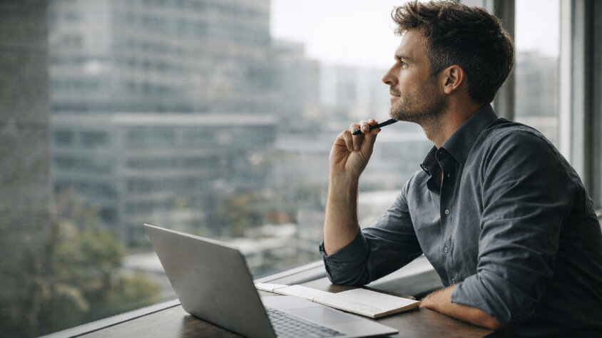 Berufstätiger Mann sitzt an einem Tisch mit Laptop und Notizbuch, blickt nachdenklich aus dem Fenster eines modernen Bürogebäudes.