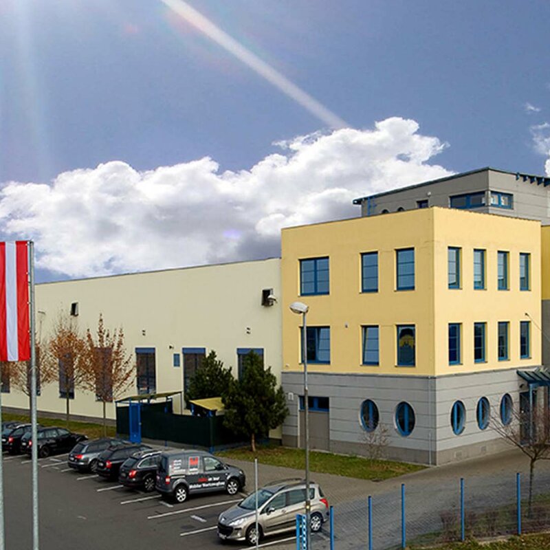 Seitenaufnahme des Gebäudes des Produktionsstandorts Olomouc aus den Gründungszeiten. Strahlendblauer Himmel mit weißen Wolken. Das Gebäude ist in gelb-grauer Farbe gehalten, und davor wehen die Flaggen der Europäischen Union, Tschechiens (CZ) und Österreichs (AT).