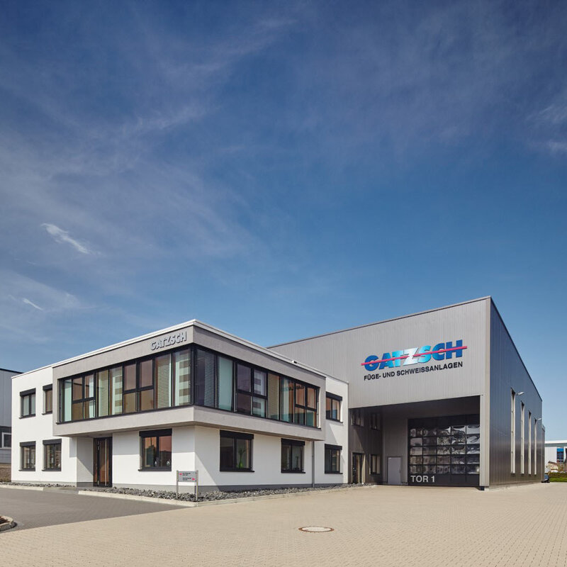 Die Außenansicht des Werkes Gatzsch Schweißtechnik GmbH zeigt ein strahlendblaues Himmel und das Verwaltungsgebäude im Fokus. Dahinter erstreckt sich die Produktionsanlage, auf der ein großes Logo mit den Worten 'Gatzsch Füge- und Schweissanlagen' deutlich sichtbar ist.