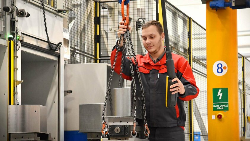 Präzises Vorrüsten in der Lohnfertigung Ein Maschinenbediener arbeitet an einem Vorrüstplatz eines automatisierten Bearbeitungszentrums für die Lohnfertigung. Er legt sorgfältig Werkzeuge und Komponenten in die Rüstplätze ein. Der Fokus liegt auf der präzisen Platzierung von Präzisionswerkzeugen in den vorgegebenen Positionen auf dem komplexen Maschinenbett. Im Hintergrund ist die industrielle Umgebung mit fortschrittlicher Technologie zu erkennen.