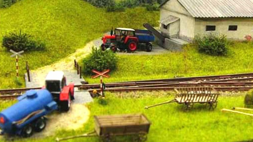 Detailreiche Modelleisenbahnlandschaft mit Gleisen, Bahnübergang, Traktoren, Anhängern und einem landwirtschaftlichen Gebäude.