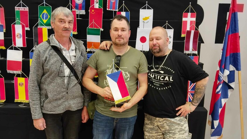 František Krumpolec steht gemeinsam mit zwei weiteren Modellbauern vor einer Ausstellung mit internationalen Flaggen.