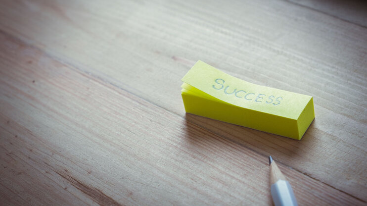 Gelber Notizzettel mit dem Wort „Success“ auf Holztisch, daneben ein Bleistift.