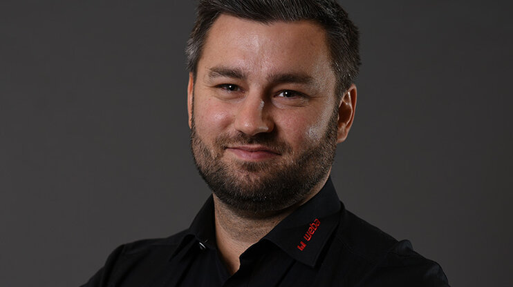 Alexander präsentiert sich freundlich und professionell: schwarzes Hemd mit rotem weba-Logo, vor grauem Hintergrund, direkt in die Kamera lächelnd.
