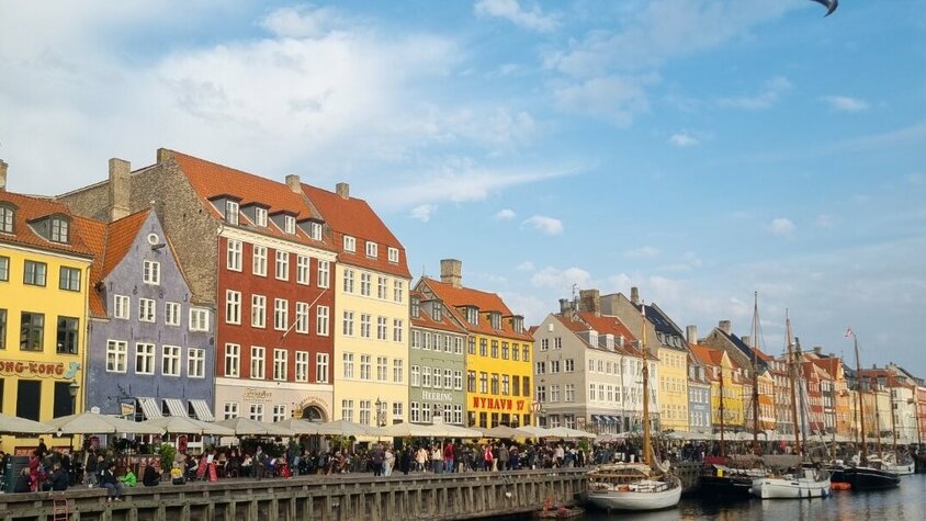 Nyhavn in Kopenhagen Nyhavn in Kopenhagen mit bunten Häusern, Booten im Wasser und vielen Menschen entlang der Promenade bei blauem Himmel.