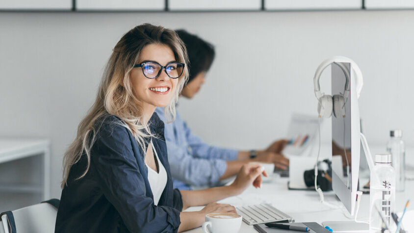 Erfolgreiche Karriereplanung: Mit den richtigen Techniken und Tools den nächsten Schritt machen Junge Frau mit Brille sitzt lächelnd im modernen Büro am Schreibtisch, arbeitet am Computer, im Hintergrund Kollegen.