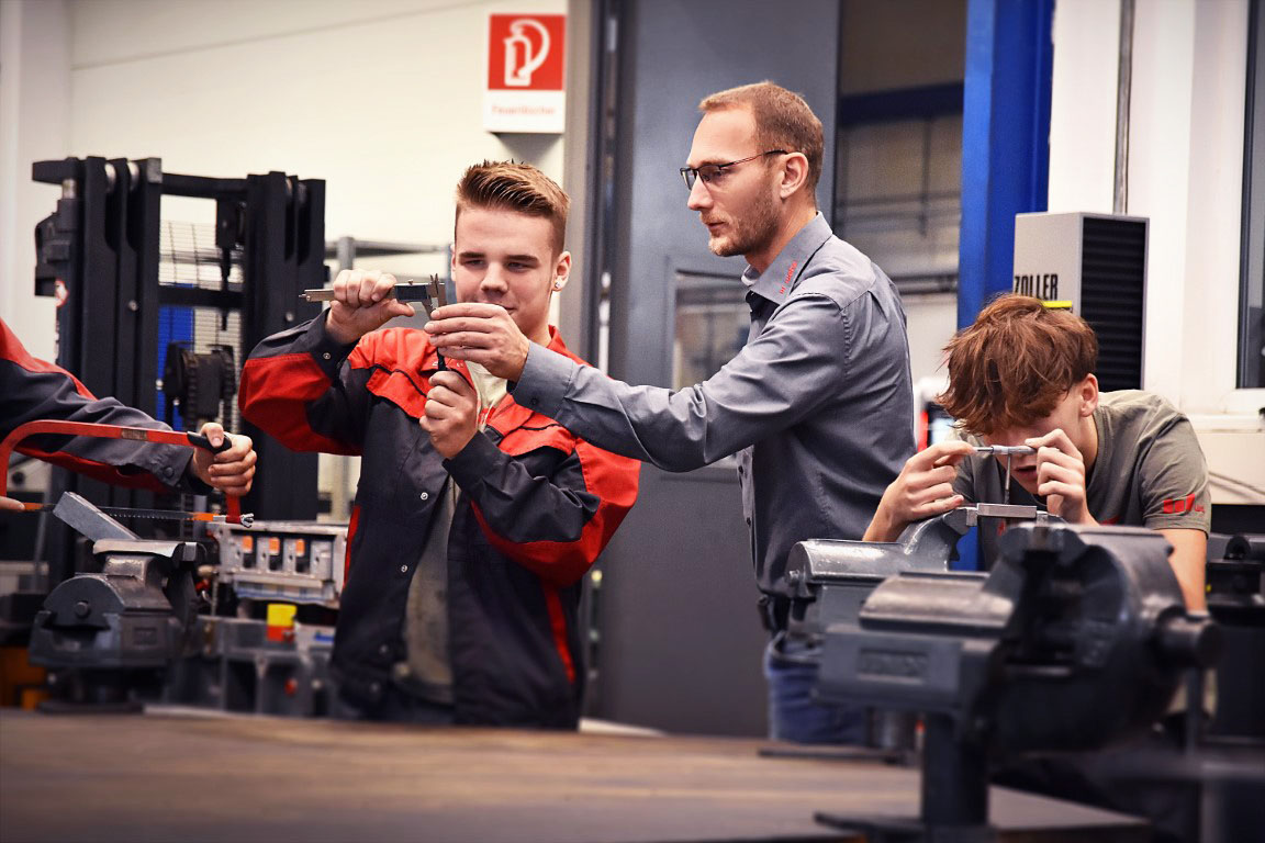 Ein Lehrling in der Werkzeugbautechnik, konzentriert beim Messen eines Stahlklotzes mit einem Messschieber, während der Ausbilder neben ihm steht und etwas erklärt und zeigt. Daneben spannt ein weiterer Lehrling aufmerksam einen Stahlklotz in einen Spannbock, was die praktische und lehrreiche Umgebung der Ausbildung verdeutlicht.