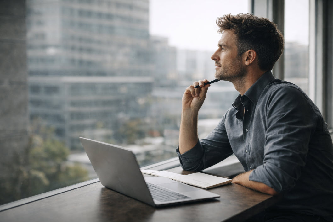 A working professional sits at a table with a laptop and notebook, thoughtfully looking out of the window of a modern office building.